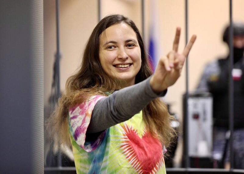 Woman in rainbow tie-dye tshirt smiles and makes a v / peace hand gesture.